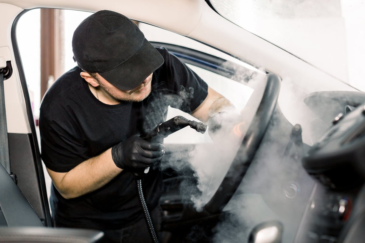 Car detailing, steam cleaning concept. Handsome man in black t-shirt and cap, worker of car wash center, cleaning car interior with hot steam cleaner. Car detailing concept.