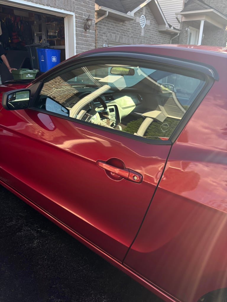 Red car with drivers side door open, showing interior with steering wheel and tan upholstery, parked in residential driveway
