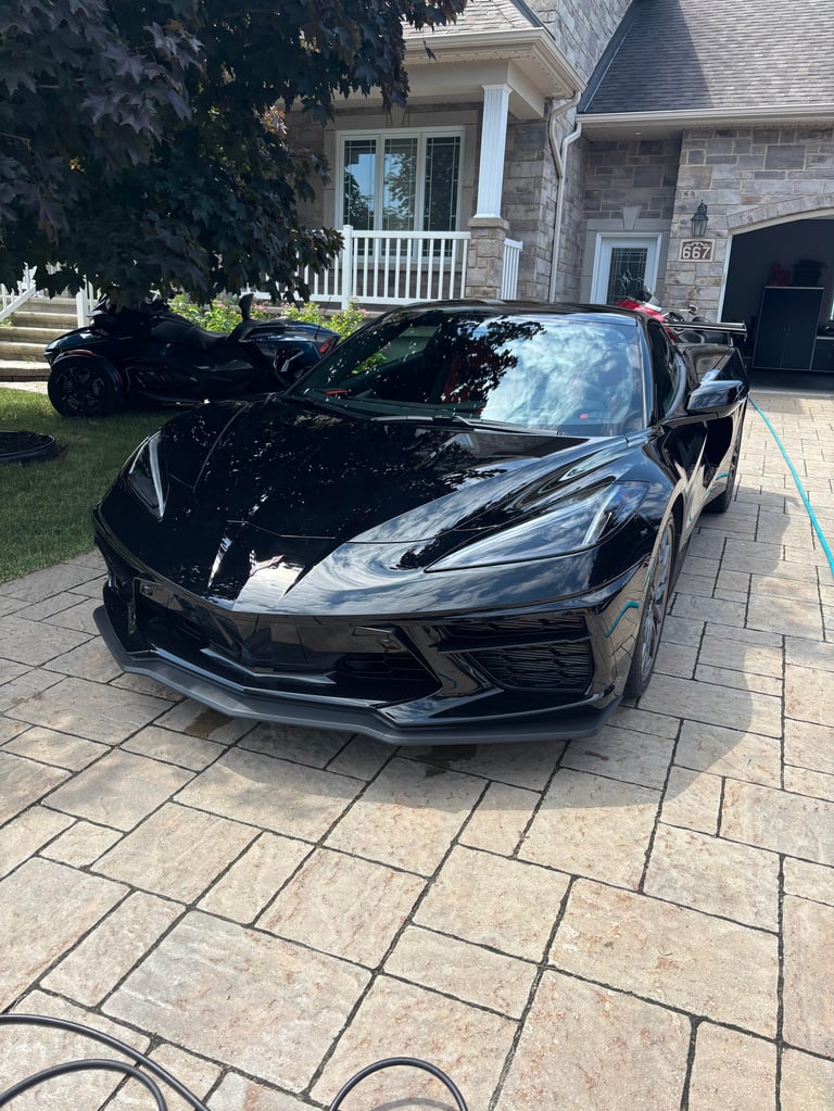 Black Corvette sports car parked on a brick driveway in front of a suburban stone house with white columns